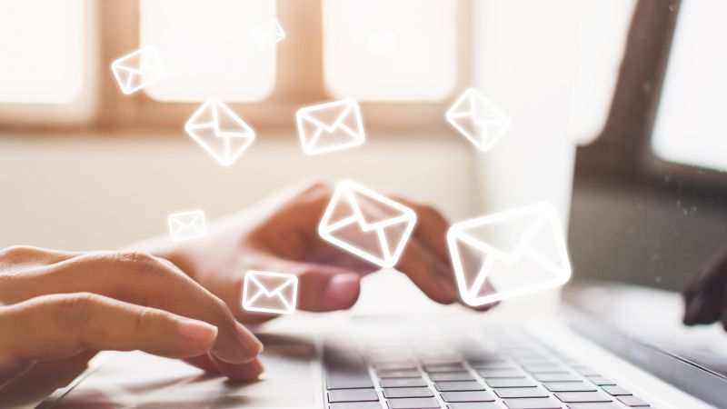 Osoba pisze na laptopie, wokół unoszą się ikony kopert symbolizujące wiadomości e-mail.