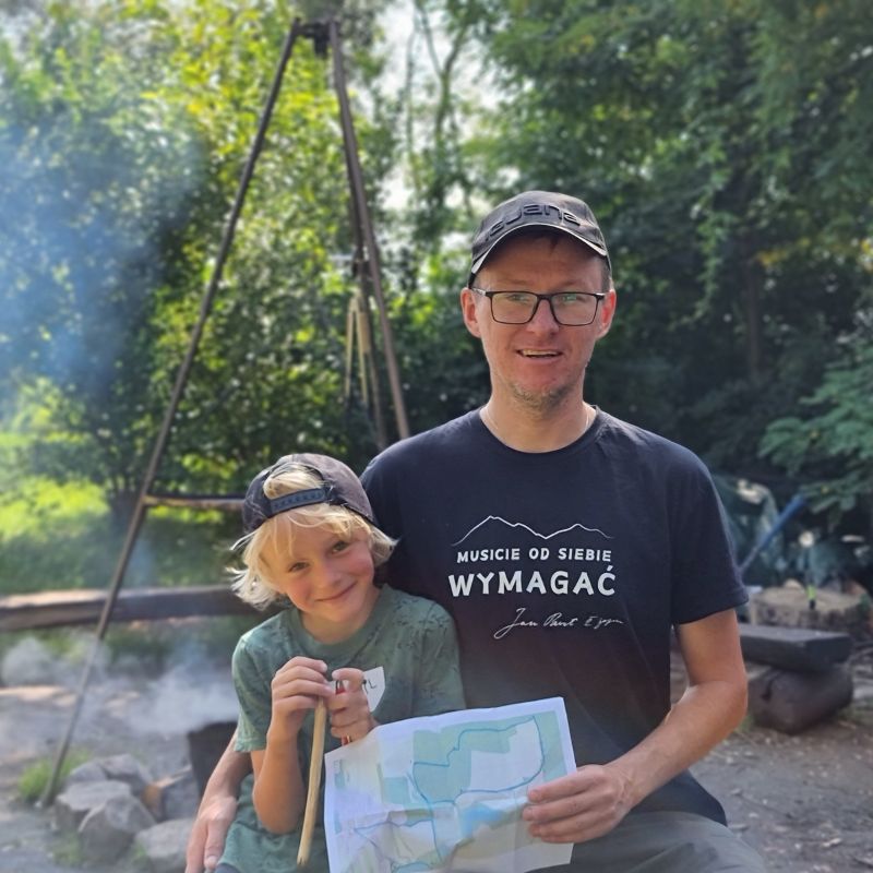 Ojciec i syn na biwaku, trzymający mapę, w otoczeniu drzew i przy ognisku.
