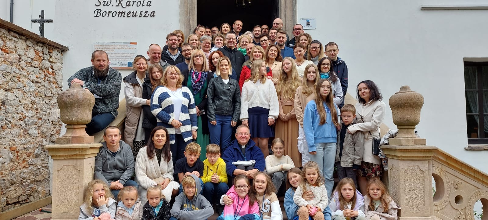 Grupa ludzi na schodach przed budynkiem z napisem Św. Karola Boromeusza, uśmiechają się do zdjęcia.