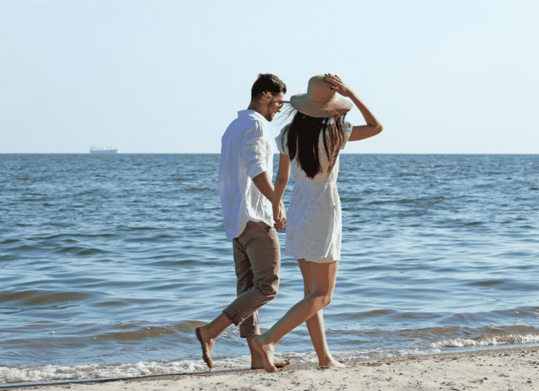 Para spacerująca boso po plaży przy słonecznej pogodzie, z widokiem na morze. Kobieta trzyma kapelusz.