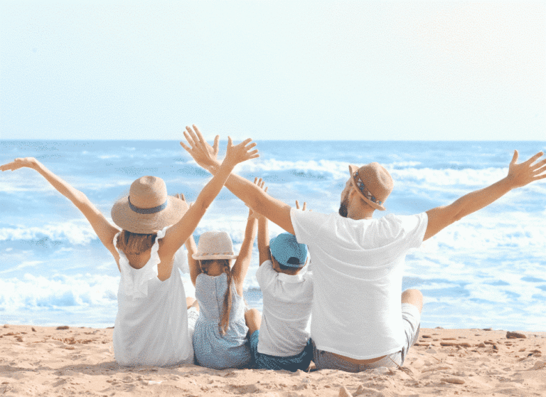 Rodzina siedzi na plaży z rękami uniesionymi do góry, patrząc na ocean w słoneczny dzień.