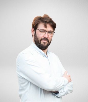 Mężczyzna w okularach z brodą stoi bokiem z założonymi rękami, ubrany w jasną koszulę, na jasnym tle.