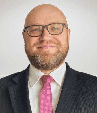 Mężczyzna w garniturze i różowym krawacie, z brodą i okularami, uśmiecha się na neutralnym tle.
