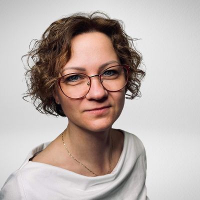 Osoba z krótkimi, kręconymi włosami i okularami, ubrana w białą bluzkę, patrzy na wprost, na jasnym tle.