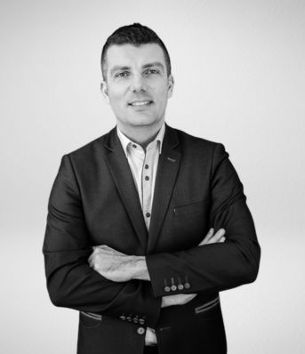 Mężczyzna w garniturze z założonymi rękami, stoi na tle białej ściany. Zdjęcie czarno-białe.