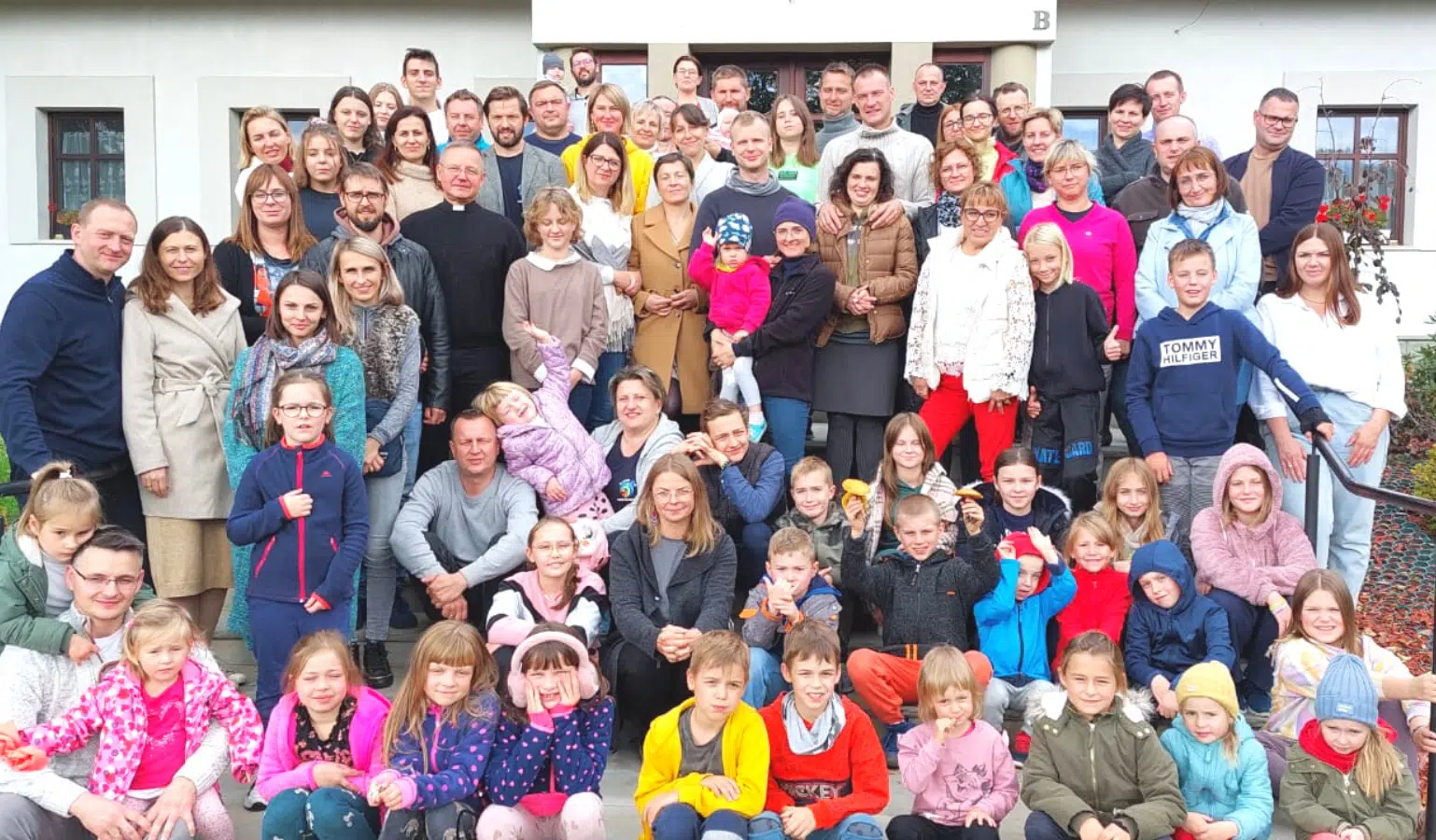 Duża grupa ludzi, dzieci i dorośli, pozują wspólnie do zdjęcia przed budynkiem. Uśmiechy i kolorowa odzież.