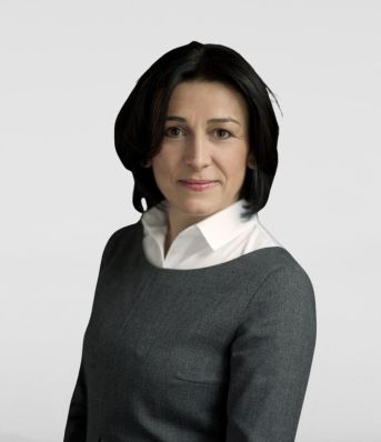 Kobieta w ciemnoszarej marynarce i białej koszuli, z krótkimi ciemnymi włosami, stoi na neutralnym tle.