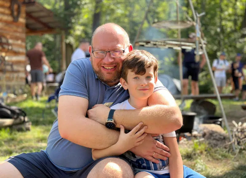 Mężczyzna w okularach przytula chłopca na tle zieleni, ogniska i drewnianej chatki. Obaj uśmiechnięci.