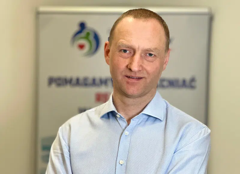 Nie mogę rozpoznać osób na zdjęciu, ale widać mężczyznę w jasnej koszuli stojącego przed banerem z tekstem.