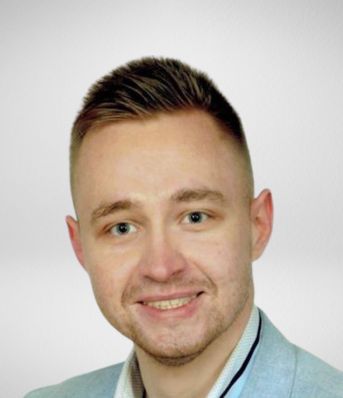 Młody mężczyzna o krótkich włosach uśmiecha się, ubrany w jasną marynarkę na białym tle.