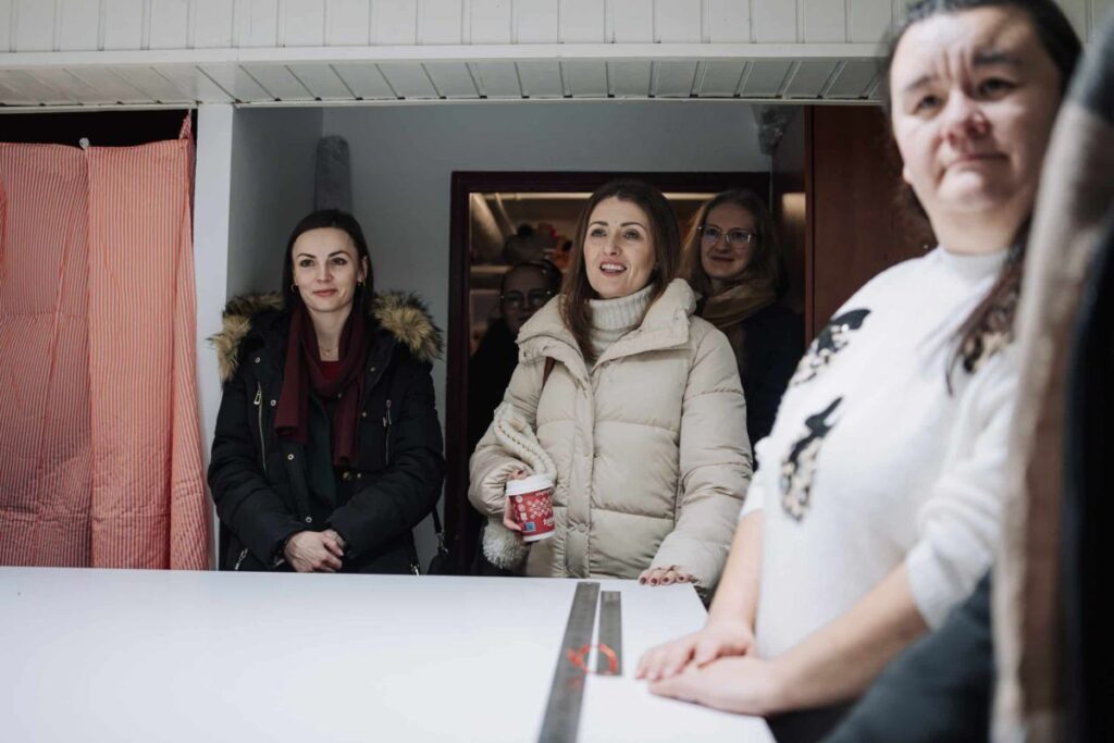 Grupa kobiet w zimowej odzieży stoi w pomieszczeniu, jedna trzyma kubek z napojem.