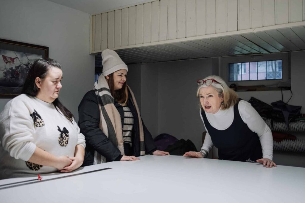 Trzy kobiety rozmawiają przy białym stole w jasnym pomieszczeniu z obrazem na ścianie. Jedna gestykuluje, pozostałe słuchają.