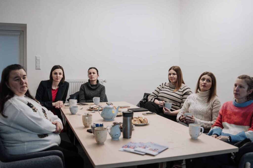 Grupa kobiet siedząca przy stole z kubkami i potrawami, uczestnicząca w spotkaniu w biurze.