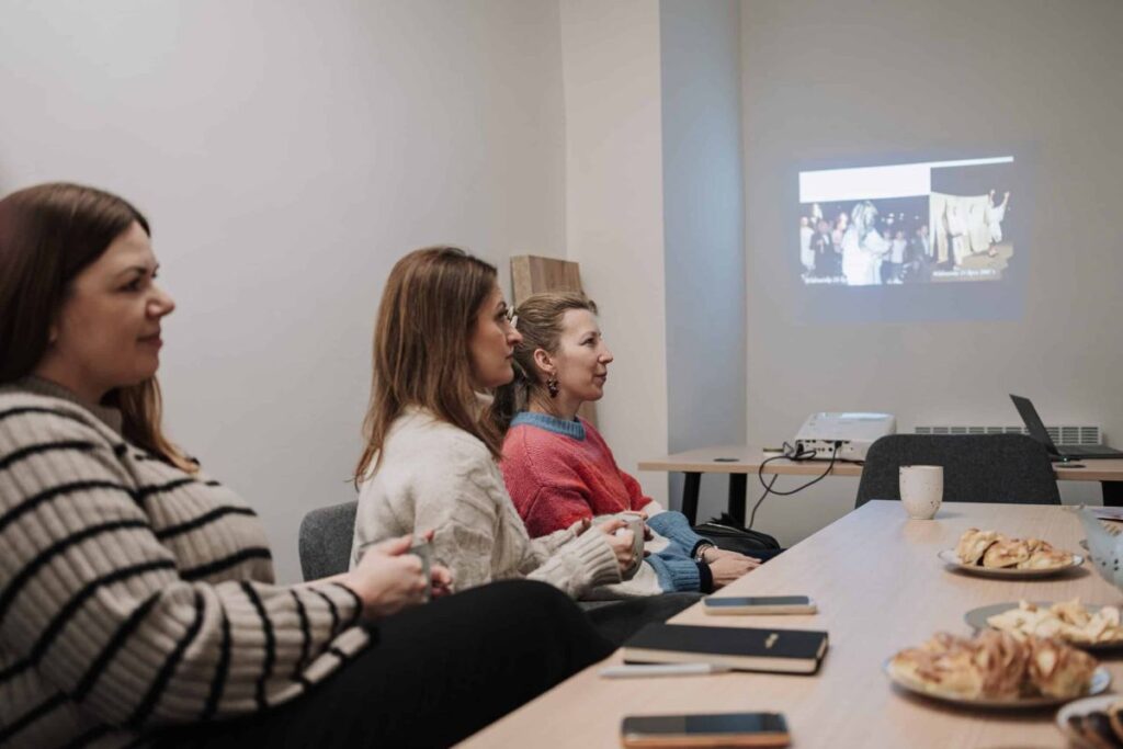 Trzy kobiety w sali konferencyjnej oglądają prezentację, na stole są notesy i talerz z przekąskami.