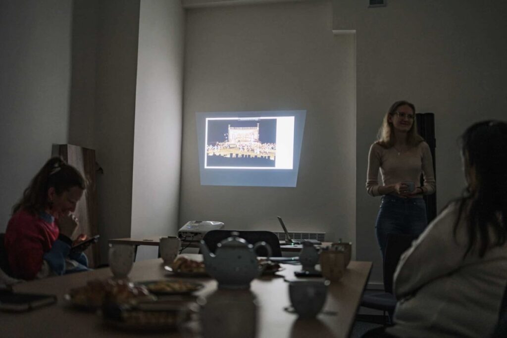 Osoby siedzą przy stole podczas prezentacji z rzutnikiem, na stole filiżanki i przekąski.