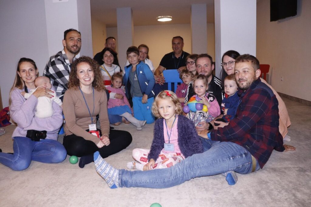 Grupa osób z dziećmi siedzi razem na dywanie w jasnym pomieszczeniu, wygląda na szczęśliwą.
