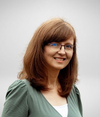 Osoba z okularami, długimi brązowymi włosami i zieloną bluzką, uśmiecha się na neutralnym tle.