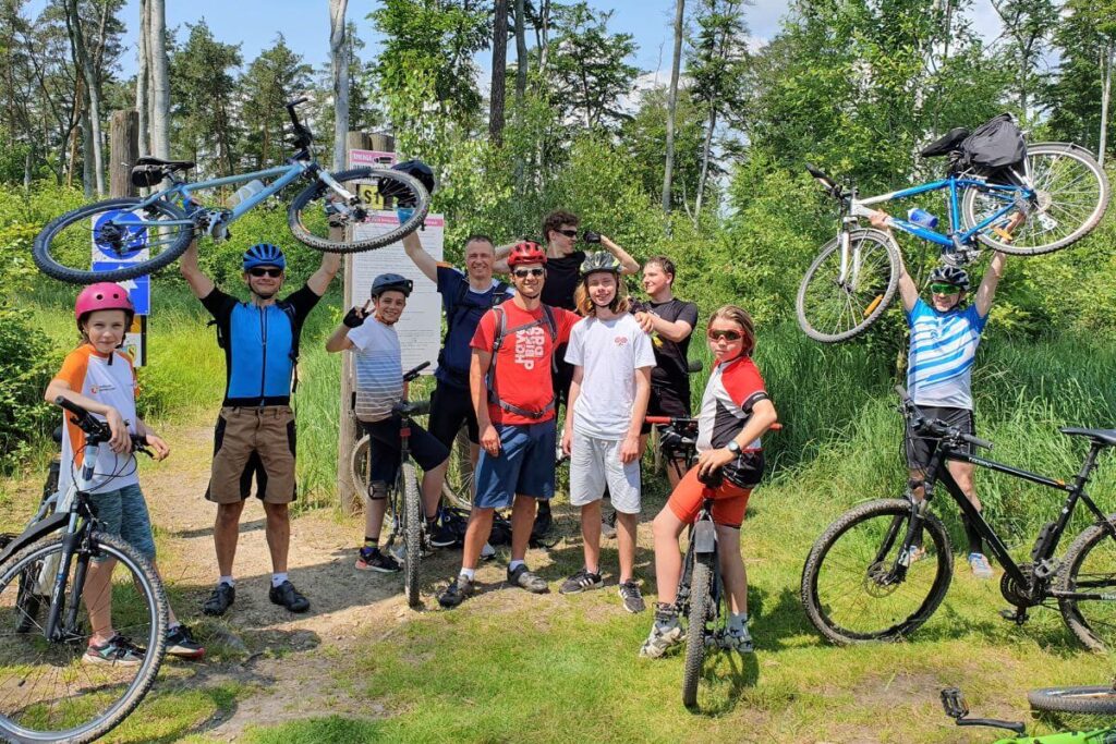 Grupa rowerzystów pozuje z rowerami na leśnej ścieżce, niektórzy podnoszą rowery w górę, w tle drzewa.