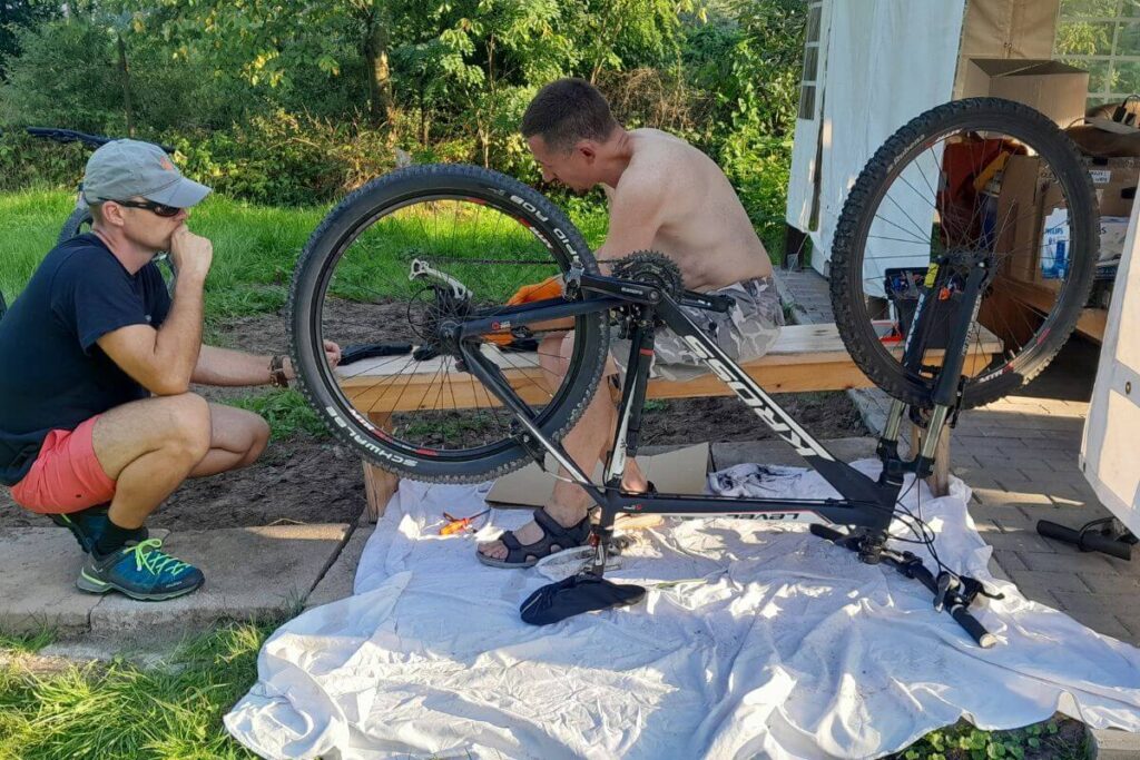 Dwóch mężczyzn naprawia rower w ogrodzie. Jeden patrzy z boku, drugi naprawia na odwróconym rowerze.