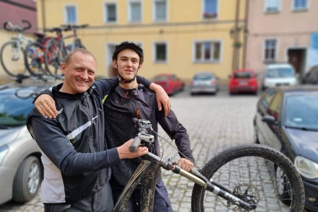 Dwóch mężczyzn w kaskach rowerowych pozuje obok roweru na tle budynku i zaparkowanych samochodów.