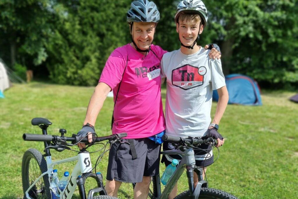Dwóch rowerzystów w kaskach i luźnych koszulkach stoi na trawie z rowerami, na tle zieleni i namiotów.