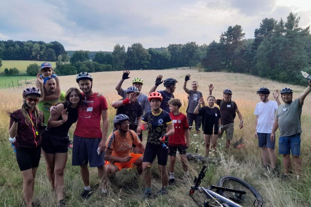 Grupa rowerzystów z uśmiechami pozuje na tle zielonego krajobrazu i błękitnego nieba.