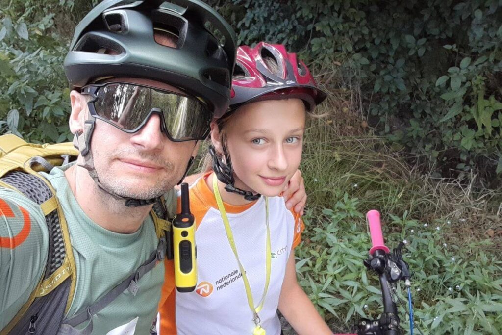 Dwóch rowerzystów w kaskach pozuje do selfie na tle zieleni. Mają na sobie sportowe ubrania.