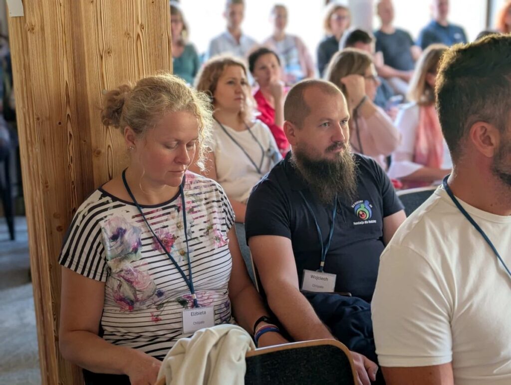 Grupa osób siedzących na spotkaniu. Kobieta i mężczyzna w pierwszym rzędzie skupieni na prezentacji.
