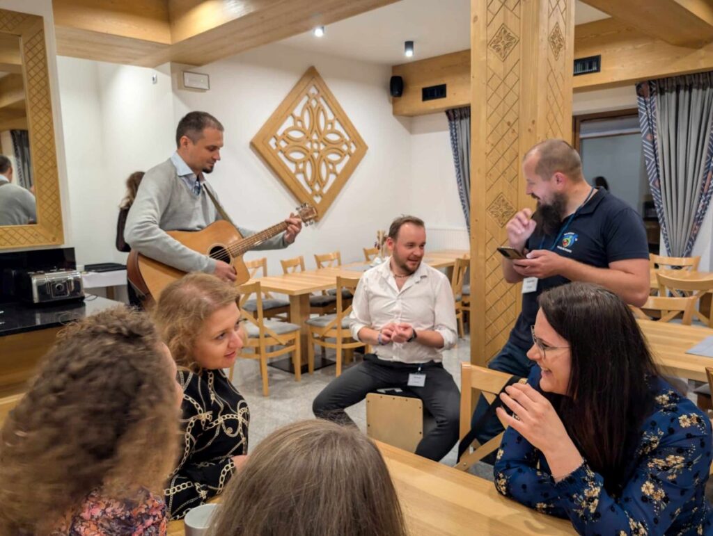Ludzie siedzą przy drewnianych stołach, jeden gra na gitarze, inni rozmawiają i śmieją się w przytulnym pokoju.