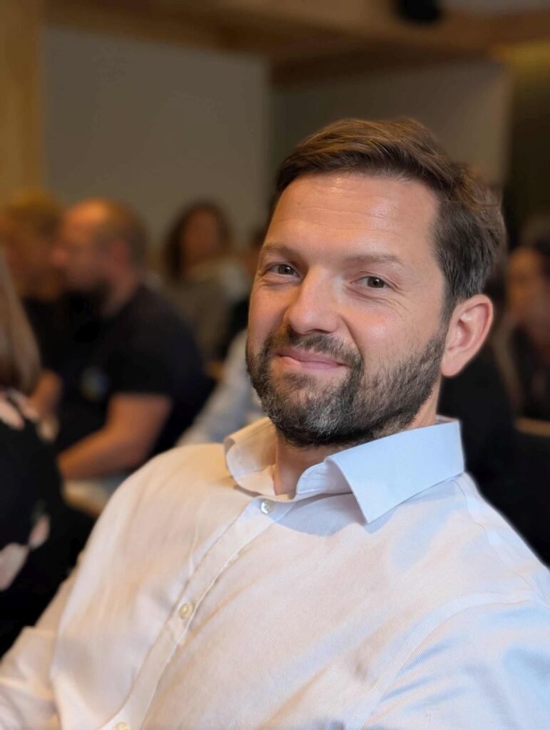 Mężczyzna w białej koszuli uśmiecha się, siedząc w tłumie na spotkaniu lub konferencji.