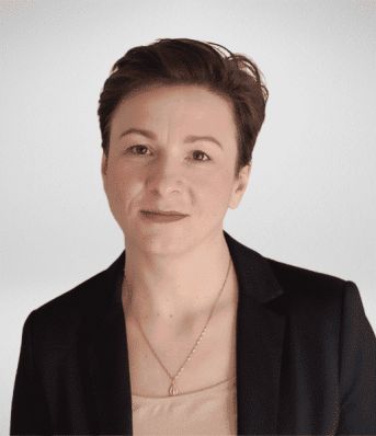 Osoba z krótkimi brązowymi włosami, w czarnej marynarce i jasnej bluzce, patrzy na wprost.