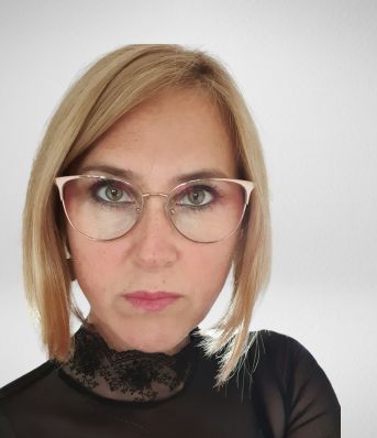 Osoba z krótkimi blond włosami i okularami patrzy w obiektyw. Ma na sobie czarną bluzkę z koronkowym wykończeniem.